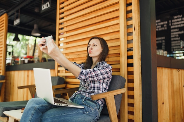 Frau im sommercafé aus holz im freien, das mit laptop-pc-computer sitzt, selfie-aufnahme auf dem handy macht und sich in der freizeit entspannt mobiles büro