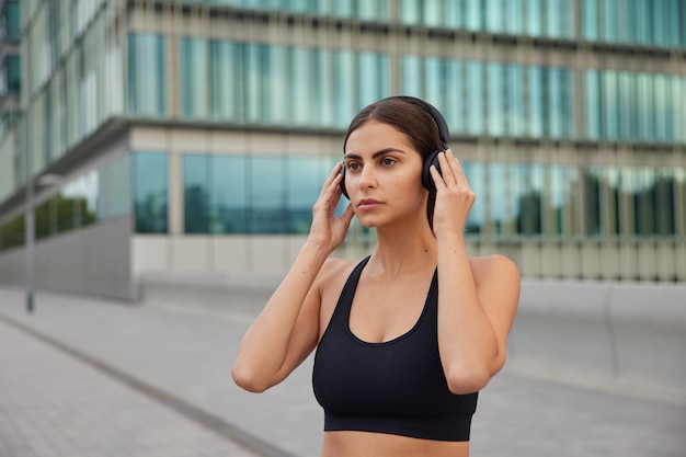 Frau im sportlichen schwarzen Top hört Musik über drahtlose Kopfhörer, die auf die Ferne fokussiert sind, geht in Sportposen in der Stadt, um im Freien zu trainieren