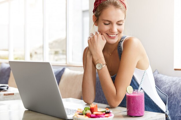 Frau im stilvollen Jeansoverall mit Laptop im Coffeeshop