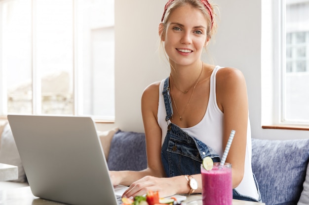 Frau im stilvollen Jeansoverall mit Laptop im Coffeeshop