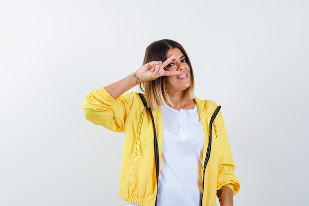 Frau im T-Shirt, Jacke, die V-Zeichen nahe Auge zeigt und fröhlich schaut, Vorderansicht.