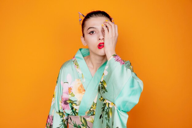 Frau im traditionellen japanischen Kimono, die schockiert ist und ein Auge mit der Hand auf Orange hält