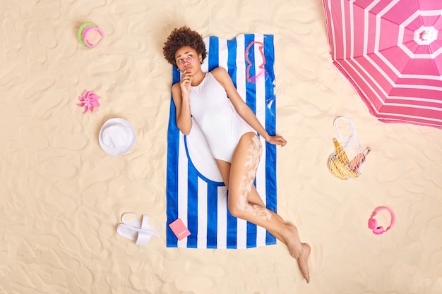 Frau im weißen Bikini posiert auf Handtuch am Sandstrand verwendet Sonnenschirm, um sich vor der Sonne zu verstecken, fühlt sich unglücklich, weil Sonnenbrand Sonnencreme auf das Gesicht aufträgt. Sommerlebensstil