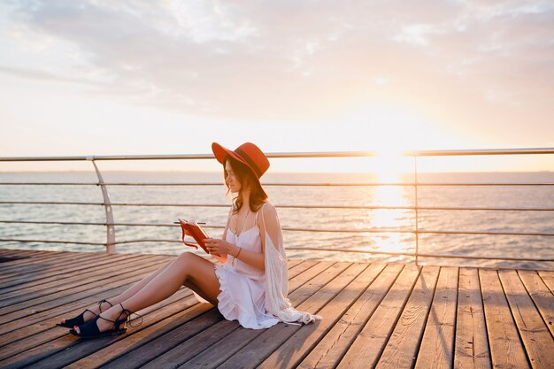 Frau im weißen Kleid, das am Meer auf Sonnenaufgang sitzt und Notizen im Tagebuchbuch in der romantischen Stimmung trägt und roten Hut trägt