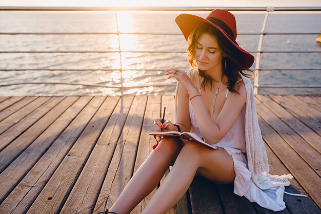 Frau im weißen Kleid, das am Meer auf Sonnenaufgang sitzt und Notizen im Tagebuchbuch in der romantischen Stimmung trägt und roten Hut trägt