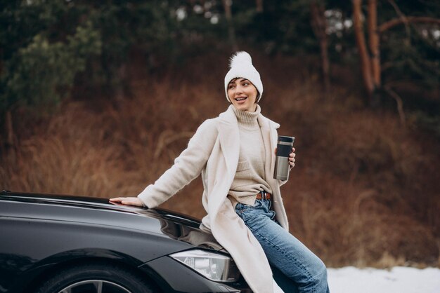 Frau im Winter sitzt auf der Motorhaube und trinkt Kaffee