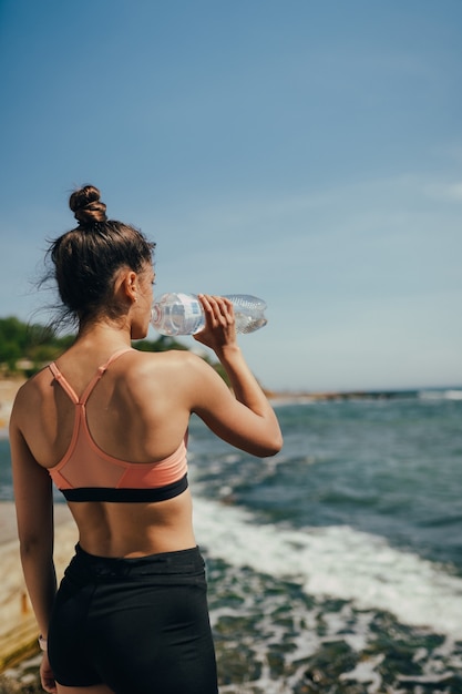 Frau im Yoga-Outfit, das frisches Wasser von der Flasche nach Übung am Strand trinkt