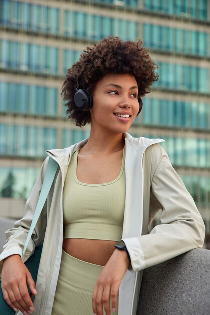 Frau in Activewear trägt Karemat schaut weg hört Musik über Kopfhörer bewundert schöne Aussicht auf die Stadt