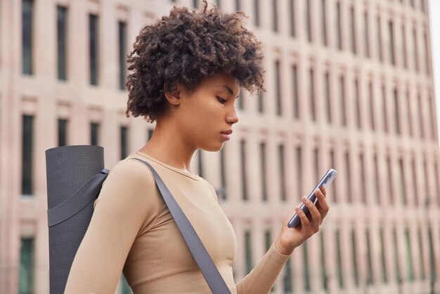 Frau in Activewear trägt Karemat zum Fitnesstraining oder Aerobic-Kurs prüft Newsfeed über Smartphone-Posen gegen Stadtgebäude