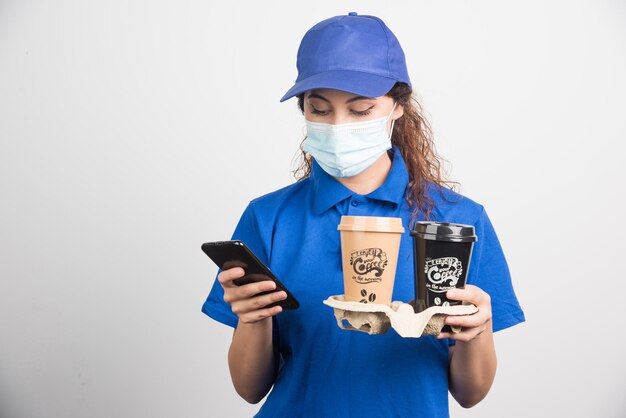 Frau in blauer Uniform mit medizinischer Maske, die auf das Telefon schaut und zwei Tassen Kaffee auf weiß hält