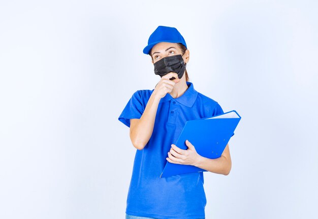 Frau in blauer Uniform und schwarzer Gesichtsmaske, die einen blauen Ordner hält und verwirrt und nachdenklich aussieht.