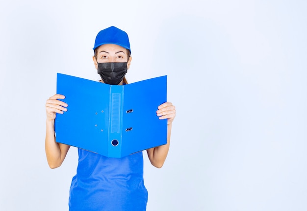 Frau in blauer Uniform und schwarzer Gesichtsmaske, die einen blauen Ordner öffnet und überprüft.