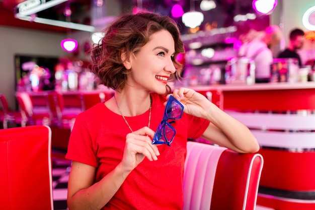 Frau in buntem Outfit im Retro-Vintage-Café der 50er Jahre, das am Tisch sitzt und rotes Hemd trägt, blaue Sonnenbrille, die Spaß in fröhlicher Stimmung hat, rotes Lippenstift-Make-up