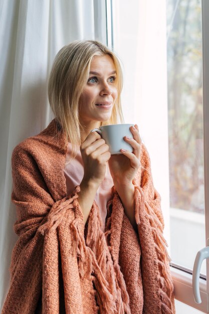 Frau in der Decke zu Hause während der Pandemie, die neben Fenster sitzt und Kaffee trinkt