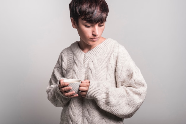 Frau in der Strickjacke, welche die Kaffeetasse steht gegen grauen Hintergrund hält