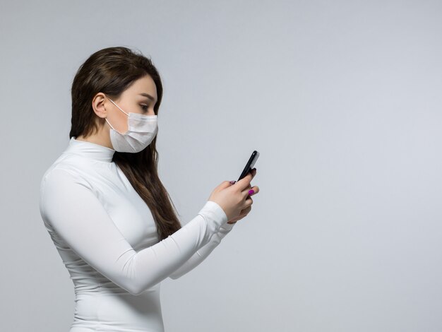 Frau in der weißen sterilen Maske, die das Telefon betrachtet