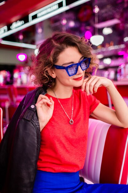 Frau in farbenfrohem Outfit im Retro-Vintage-Café der 50er Jahre, das am Tisch mit Jacke, blauem Rock und rotem Hemd, Sonnenbrille sitzt