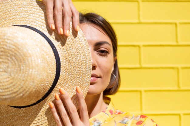 Frau in gelbem Sommerkleid und Hut auf gelber Backsteinmauer ruhig und positiv, genießt sonnige Sommertage