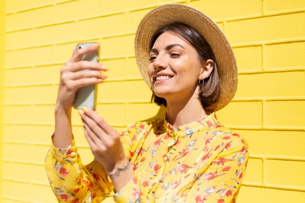 Frau in gelbem Sommerkleid und Hut auf gelber Backsteinmauer ruhig und positiv haltendes Mobiltelefon