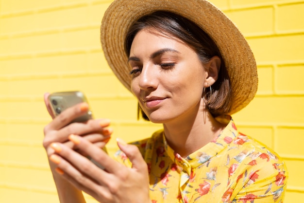 Kostenloses Foto frau in gelbem sommerkleid und hut auf gelber backsteinmauer ruhig und positiv haltendes mobiltelefon