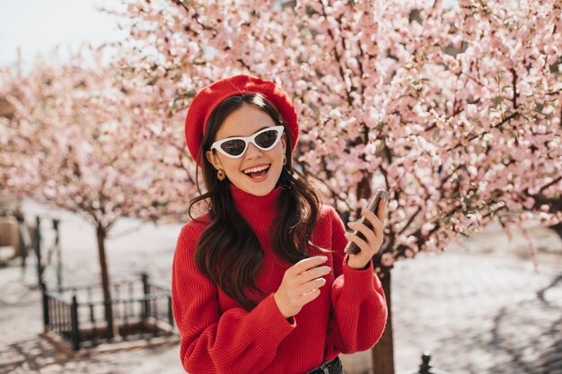 Frau in Hochstimmung wirft mit Telefon nahe Kirschblüten auf. Porträt der Dame in der roten Baskenmütze, im Kaschmirpullover und in den weißen Brillen im Garten im Frühjahr