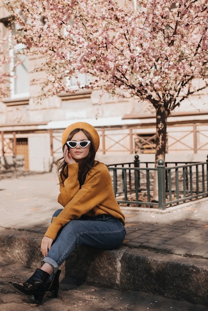 Frau in Jeans und Pullover ruht auf Bordstein vor dem Hintergrund von Sakura. Porträt der Dame im stilvollen Outfit im Pariser Stil, das Frühlingswetter genießt