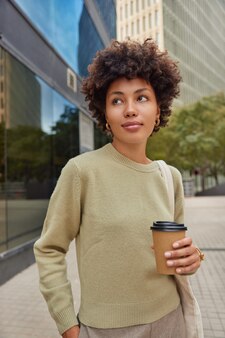 Frau in lässigem outfit spaziert im freien trinkt kaffee zum mitnehmen hält die hand in der tasche konzentriert in distanzposen gegen die städtische umgebung