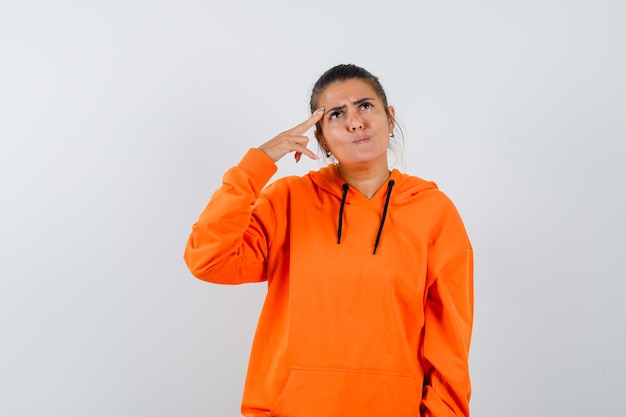 Frau in orangefarbenem Hoodie, die Finger an den Schläfen hält und nachdenklich aussieht