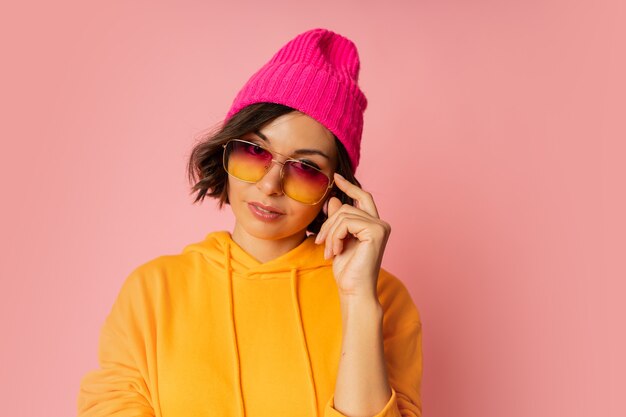 Frau in rosa Hut und orangefarbenem Hoodie posiert auf Rosa. Stilvolle Sonnenbrille.