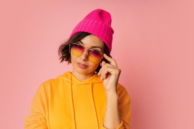 Frau in rosa Hut und orangefarbenem Hoodie posiert auf Rosa. Stilvolle Sonnenbrille.