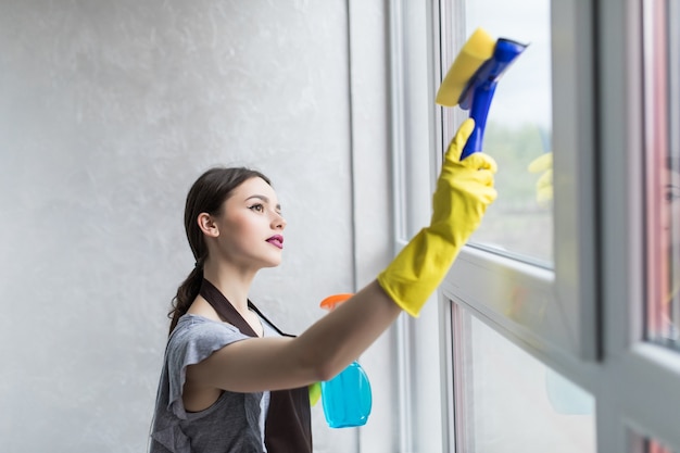 Frau in Schutzhandschuhen lächelt und wischt Staub mit einem Spray und einem Staubtuch ab, während sie ihr Haus putzt, Nahaufnahme close