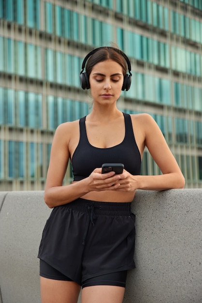 Frau in Sportbekleidung lädt Songs für das Training in die Playlist herunter liest Nachrichten über den letzten Wettbewerb durchsucht die Website überprüft die Benachrichtigung von der Sportanwendung stellt sich gegen das Glasgebäude