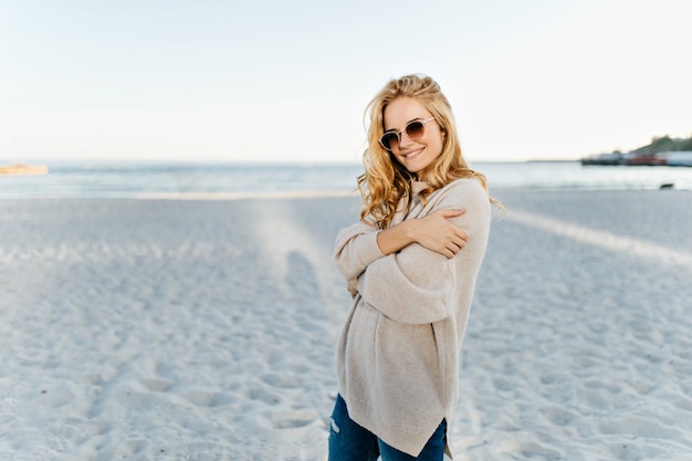 Frau in übergroßem Pullover und Jeans posiert in guter Stimmung am Strand gegen Meer.