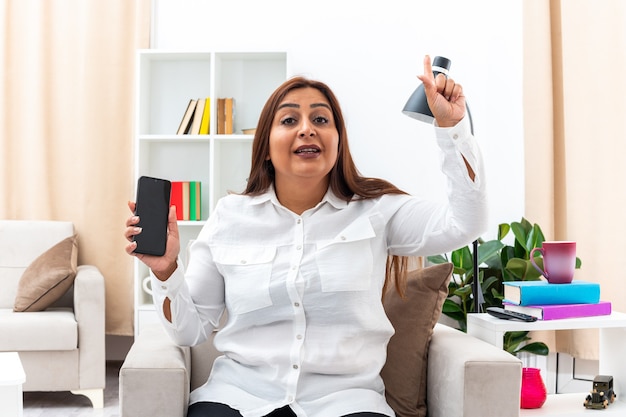 Frau in weißem Hemd und schwarzer Hose mit Smartphone lächelnd mit Zeigefinger mit neuer Idee auf dem Stuhl im hellen Wohnzimmer sitzend