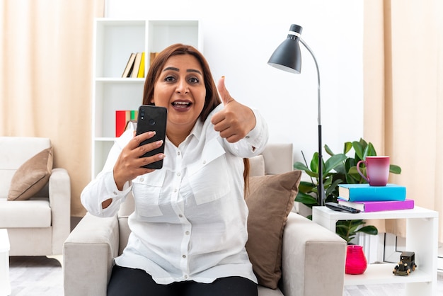 Frau in weißem Hemd und schwarzer Hose mit Smartphone zeigt Daumen hoch glücklich und positiv lächelnd auf dem Stuhl im hellen Wohnzimmer sitzend