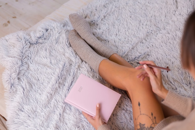 Frau in Wollsocke und -pullover mit rosa Notizbuchzeichen 2021, großes Tattoo auf Hüfte. Frau sitzen zu Hause im Schlafzimmer auf dem Bett.