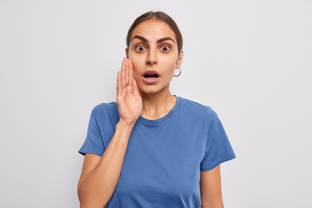 Frau keucht vor Verwunderung hält die Hand in der Nähe des Gesichts Mund geöffnet verbreitet Gerüchte flüstert etwas mit schockiertem Gesichtsausdruck gekleidet in einem lässigen blauen T-Shirt auf Weiß