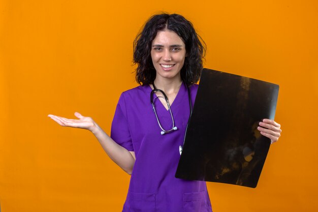 Frau Krankenschwester in der medizinischen Uniform und mit Stethoskop, das Röntgenstrahl hält und mit der Hand zur Seite zeigt, die mit glücklichem Gesicht lächelt, das auf lokalisiertem Gelb steht