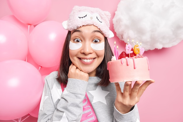 Frau lächelt fröhlich feiert Geburtstag in häuslicher Atmosphäre trägt Schlafmaske und Pyjama hält leckeren Kuchen