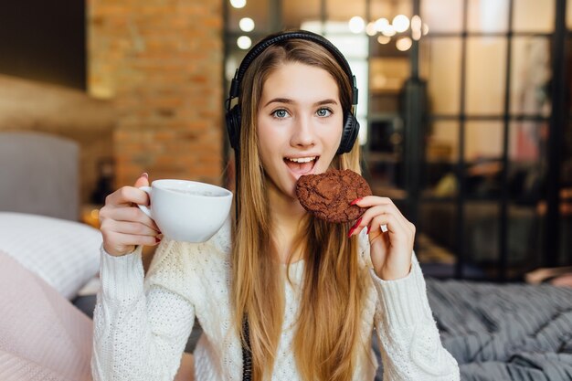 Frau lächelt, während sie Kuchen isst, Kaffee trinkt und Kopfhörer trägt, die mit dem Tablet-Gadget verbunden sind