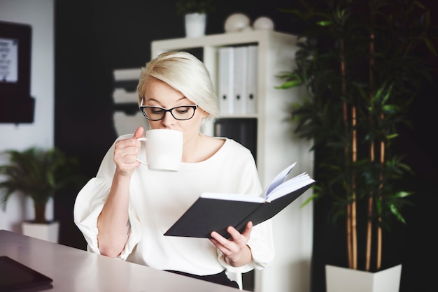 Frau liest Buch und trinkt Kaffee an ihrem Schreibtisch