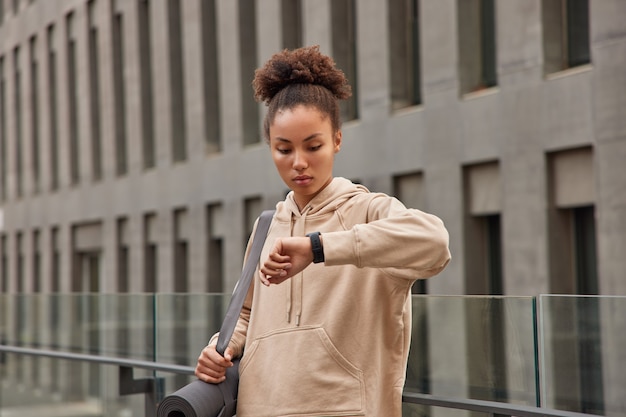 Frau macht Pause, nachdem Fitnessaktivitäten die Ergebnisse auf der Smartwatch überprüft haben, trägt gerollte Karemat in Hoodie-Ständern gegen modernes Gebäude