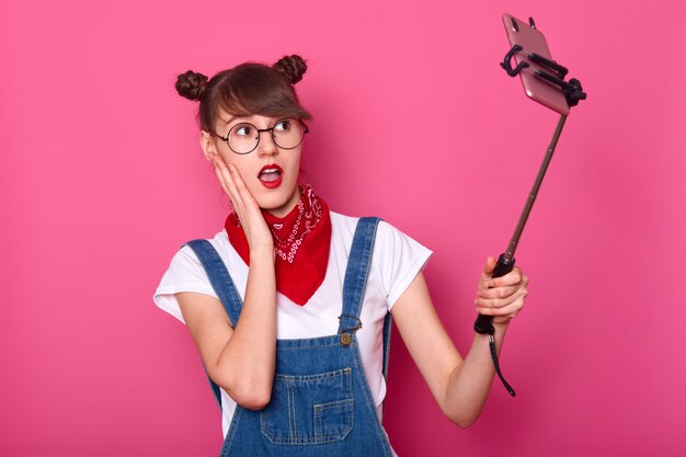 Frau macht Selfie, benutzt Selfie-Stick, posiert, macht Foto für ihr soziales Netzwerk, sieht überrascht aus, steht mit offenem Mund, hält die Hand auf der Wange. Teen Konzept.