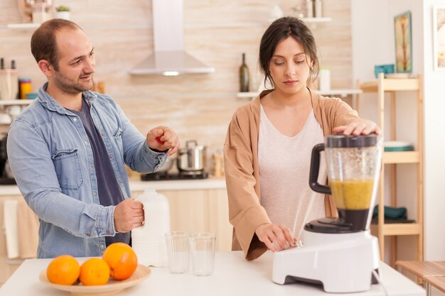 Frau mischt Früchte in Smoothie Maker, während Ehemann Milchflasche öffnet. Gesunder, unbeschwerter und fröhlicher Lebensstil, Ernährung und Frühstückszubereitung am gemütlichen sonnigen Morgen