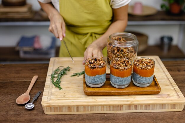 Frau mit bunten gesunden süßen Wüsten-Chia-Puddings aus Mandelmilch, blauem Spirulina-Extrakt, Chiasamen, Pappaya-Mango-Marmelade und hausgemachtem Müsli. Auf Holztisch in der Küche zu Hause.