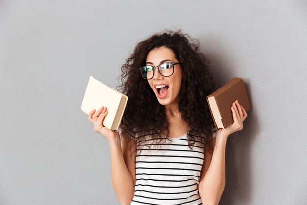 Frau mit dem braunen gelockten Haar, das Student in der Universität aufwirft mit interessanten Büchern in den Händen ist, welche die Bildung genießen, die über grauer Wand lokalisiert wird