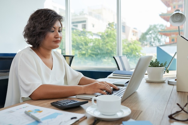 Frau mit dem kurzen gewellten Haar, das am Schreibtisch im Büro sitzt und an Laptop arbeitet