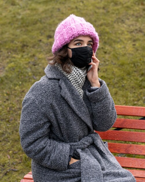 Frau mit der medizinischen Maske in der Stadt, die über das Telefon auf Bank unterhält