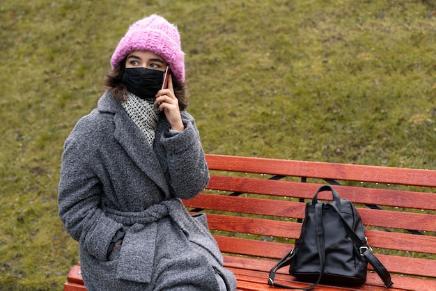 Kostenloses Foto frau mit der medizinischen maske in der stadt, die über das telefon auf bank unterhält