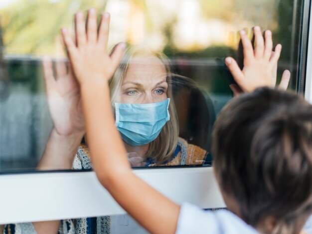 Frau mit der medizinischen Maske zu Hause, die Neffen durch das Fenster salutiert